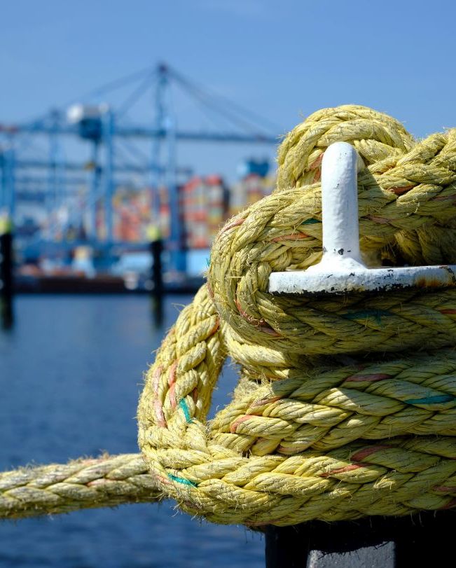 A iron bollard with a tied rope on a quay in the Port of Rotterdam in the Netherlands. In the background, slightly out of focus, is the industrial area of the Maasvlakte near Rotterdam.